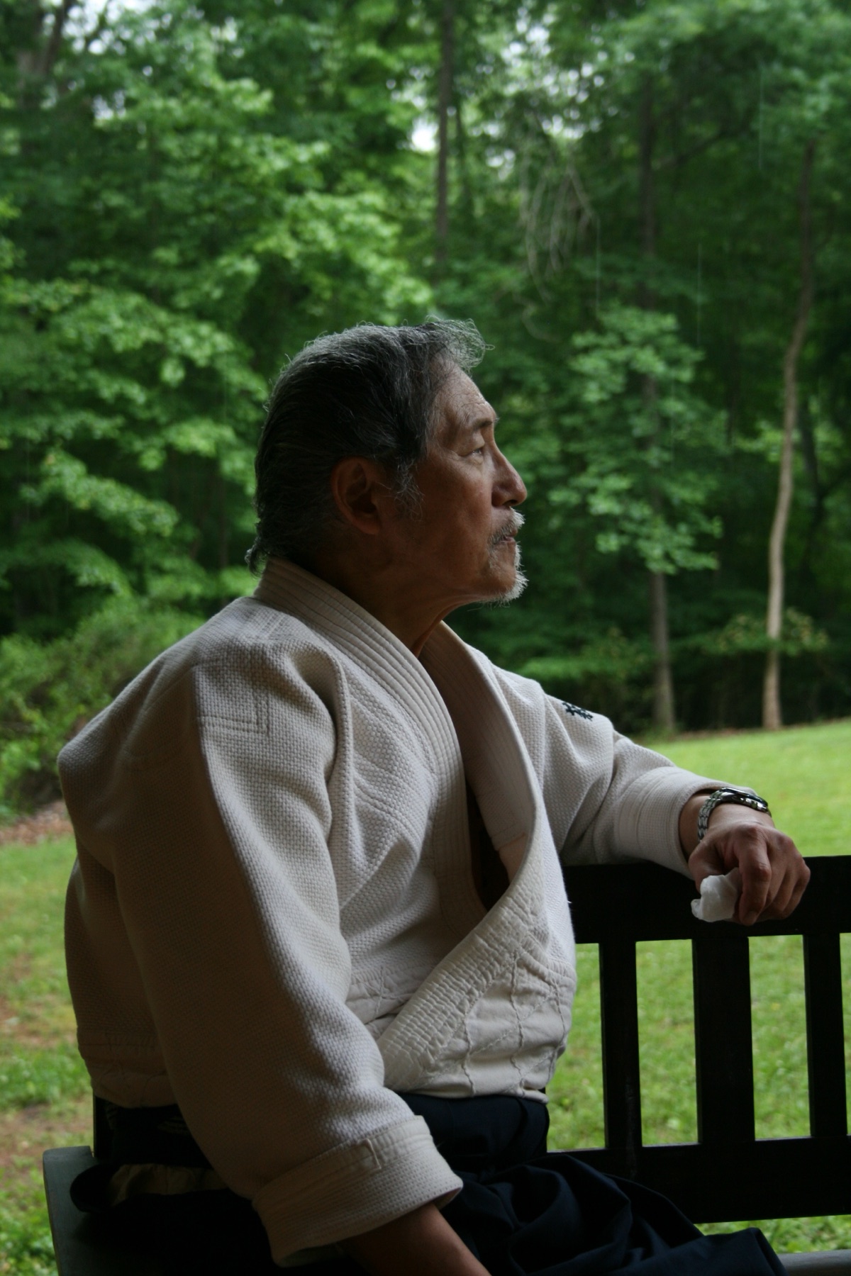 Edward Hagihara in a white keikogi and dark hakama, seated on a wooden bench at the edge of a wood, looking into the distance